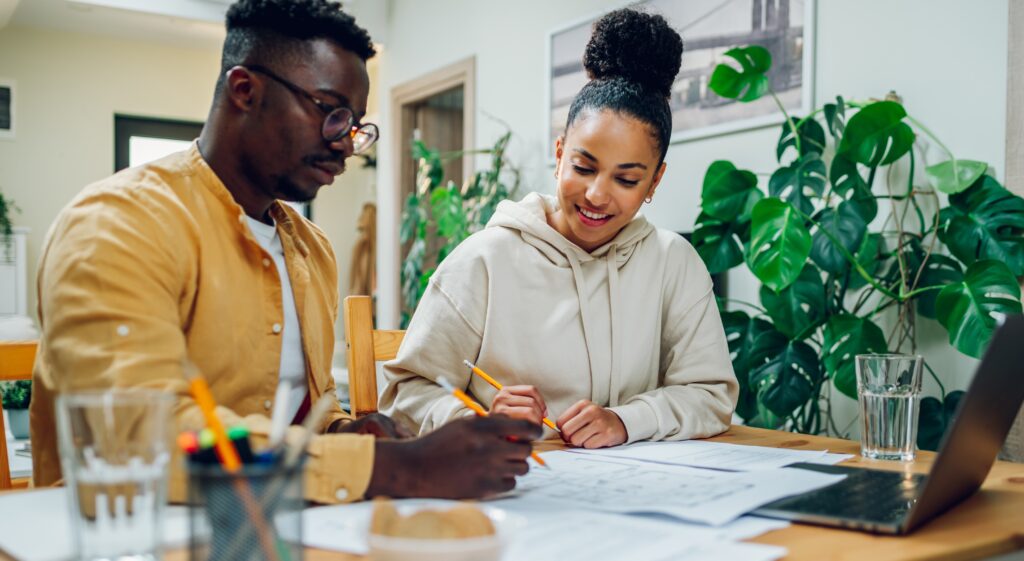 A man and woman sit at a table reviewing documents together with pencils in hand, smiling and discussing plans. A laptop, glasses of water, and office supplies are on the table, with houseplants in the background.