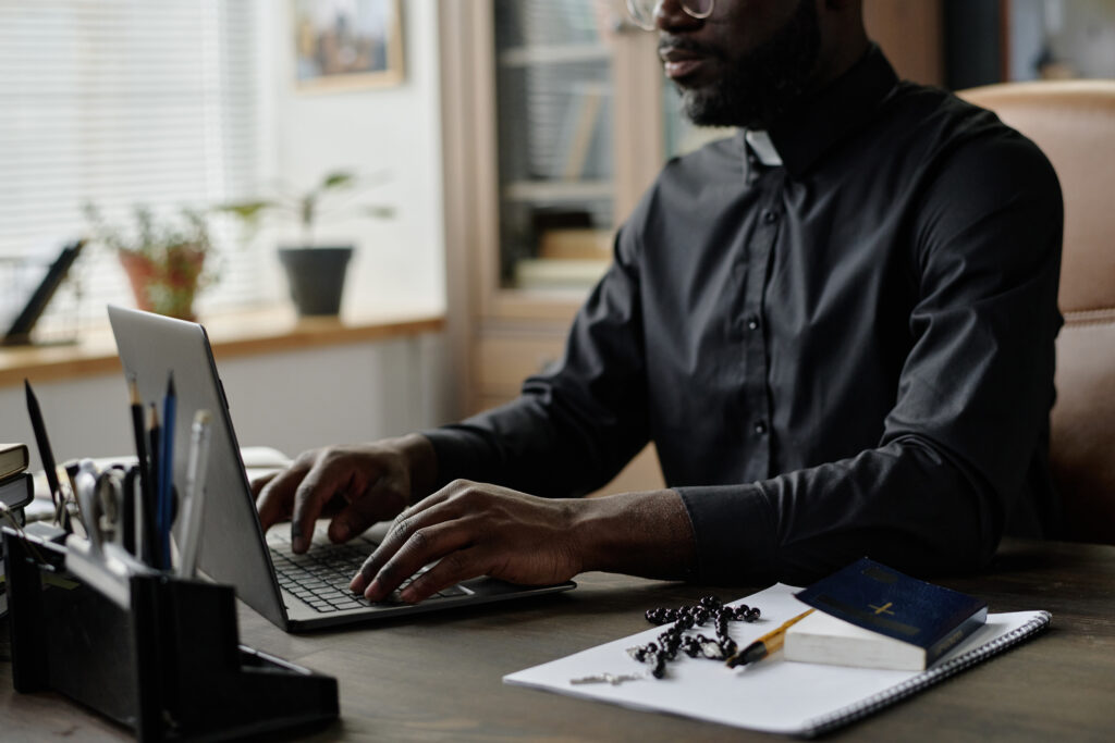 African American pastor typing on laptops keyboard while sitting at table with rosary, bible and office supplies on top
