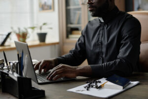 African American pastor typing on laptops keyboard while sitting at table with rosary, bible and office supplies on top