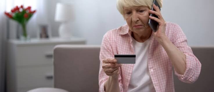 An older adult seated indoors holds a credit card in one hand and a smartphone to the ear with the other hand. The scene shows a home setting with a blurred background, including a table, a lamp, and a plant, while the person looks down at the card during the phone call.