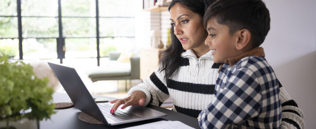 A person and a child sit together at a table using a laptop in a bright home office or living space. Papers are spread on the table, and large windows and shelving are visible in the background.