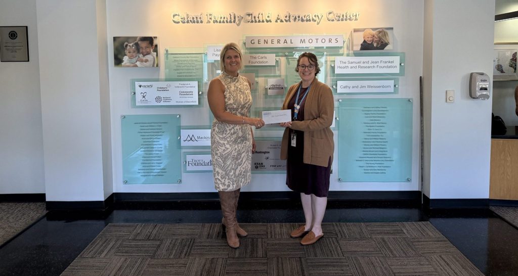 Two individuals stand in the lobby of CARE House of Oakland County's Galan Family Child Advocacy Center, holding a donation check together in front of a donor recognition wall.